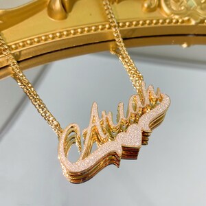 K&ouml;nnte beinhalten: Eine goldene Kettenkette mit einem mehrschichtigen Namensschild-Anh&auml;nger, auf dem "Ava" mit einem kleinen Herz-Anh&auml;nger steht. Das Namensschild ist mit glitzerndem Glitzer bedeckt.