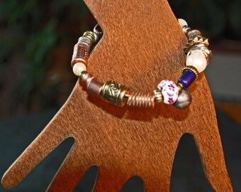Puede incluir: Una pulsera de cuentas decorativa con una mezcla de cuentas de metal, perlas y cerámica. La pulsera presenta cuentas plateadas, doradas, cobrizas, blancas, azules y moradas. La pulsera se exhibe en un soporte de madera con forma de mano.
