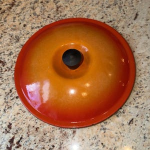 May include: A round, orange and red enameled pot lid with a black knob. The lid has a glossy finish and a gradient color scheme, transitioning from red at the edges to orange in the center. A kitchenware item.