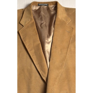 May include: A close-up of a tan corduroy blazer. The blazer has a notched lapel, a two-button closure, and a single chest pocket. The interior lining is a light tan color. The blazer appears to be in good condition.