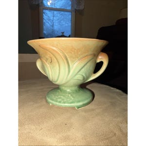 May include: A vintage ceramic vase with a two-tone color scheme. The top portion is a warm, earthy orange, transitioning to a soft green at the base. The vase features a pedestal base, two curved handles, and a flared rim. The design includes embossed leaf-like patterns.