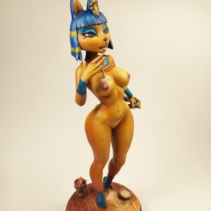 May include: A stylized figurine of a feline-humanoid figure, painted in shades of yellow and blue. The figure is posed with one arm raised, and stands on a circular base with hieroglyphic patterns. The figurine has a cat-like face and is adorned with jewelry.