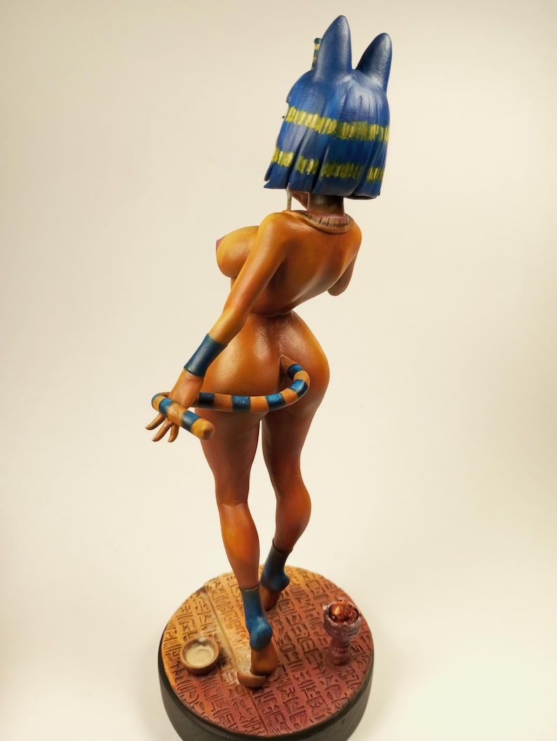 May include: A detailed figurine of a nude figure with a blue and yellow headdress and matching armbands and anklets. The figure has a striped tail and is standing on a round base with hieroglyphs.