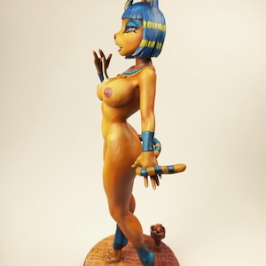May include: A figurine of a woman with a cat-like head, wearing blue and gold accents. The figure is nude, with a detailed design, and stands on a round base. The figurine is approximately 8 inches tall.