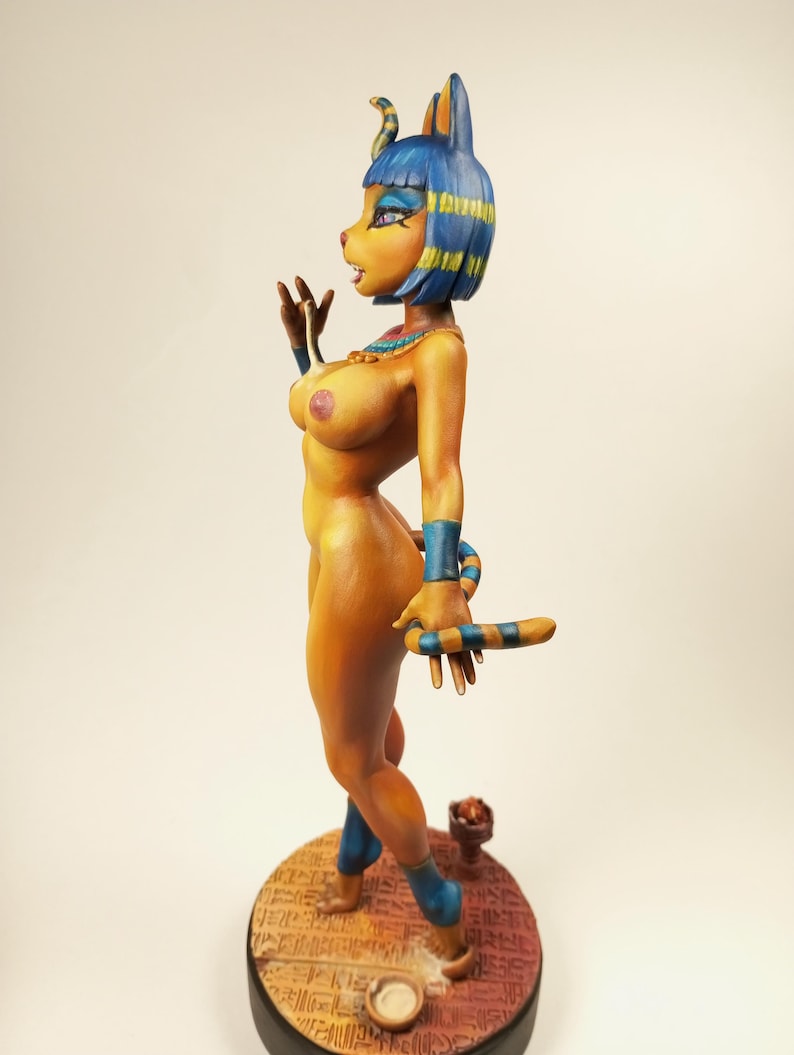 May include: A figurine of a woman with a cat-like head, wearing blue and gold accents. The figure is nude, with a detailed design, and stands on a round base. The figurine is approximately 8 inches tall.