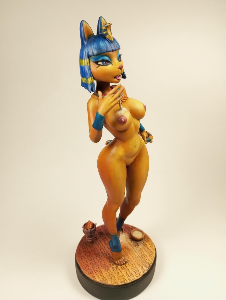 May include: A detailed figurine of a cat-like character with blue hair and accents, wearing blue armbands and sandals. The figure is posed on a round base with hieroglyphic-style carvings, and is painted in shades of yellow, orange, and blue.