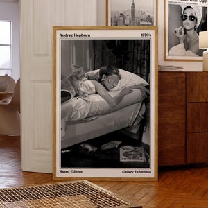 May include: A framed black and white print featuring Audrey Hepburn in bed with a cat, labeled "Audrey Hepburn 1970's Retro Edition." Two other framed prints are visible, one with the text "The Travel Times NEW YORK" and another with "The New York Trends Better Late Than Ugly."