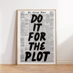 May include: Framed print featuring a vintage newspaper background with the bold text "DO IT FOR THE PLOT" in black. The print is in a wooden frame and is displayed against a white wall and wooden floor.
