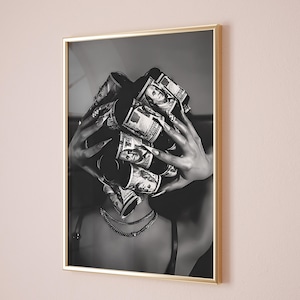 May include: Black and white framed art print. The image features a person with rolls of US hundred dollar bills covering their head. The person is wearing a black top and multiple gold necklaces. The frame is gold.