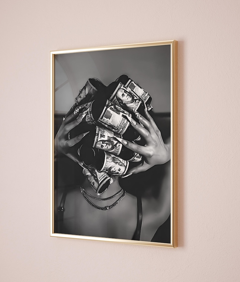 May include: Black and white framed art print. The image features a person with rolls of US hundred dollar bills covering their head. The person is wearing a black top and multiple gold necklaces. The frame is gold.