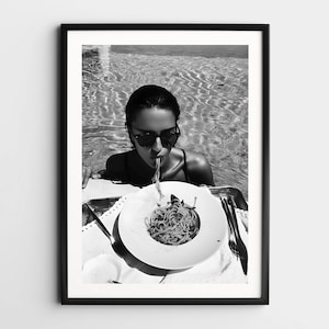May include: A black and white photograph of a person in a swimming pool eating spaghetti. The person is wearing sunglasses and is sitting at a table with a plate of spaghetti.