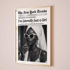 May include: A framed black and white print featuring a woman applying lipstick, wearing white sunglasses, and a towel on her head. The print is a mock newspaper with the headline "I'm Literally Just a Girl."