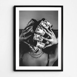 May include: Black and white framed print featuring a person with long nails holding rolled-up US dollar bills around their head. The person is wearing a necklace and a black top. The image is a close-up shot.