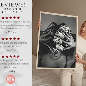 May include: Black and white print of a person with rolled-up US dollar bills covering their head. The print is held by a person wearing a white top and beige pants. The print is in a wooden frame.