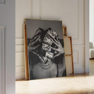 May include: Black and white framed art print featuring a person with hands holding rolled-up US dollar bills in front of their face. The print has a gold frame and is leaning against a wall.