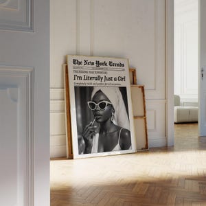May include: A framed black and white print of a woman wearing sunglasses and a towel, holding a lipstick. The print is designed to look like a newspaper article titled "The New York Trends" with the headline "I'm Literally Just a Girl."