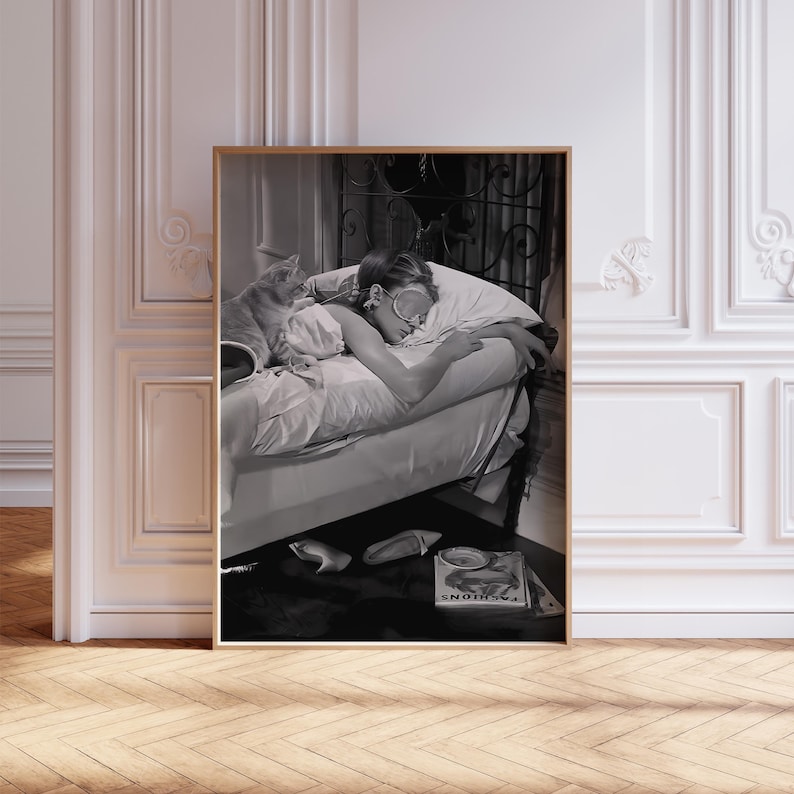 May include: Black and white photo of a woman sleeping in bed with a cat. The woman is wearing a sleep mask and is lying on her side. The cat is curled up on the bed next to her. The photo is framed in a wooden frame.