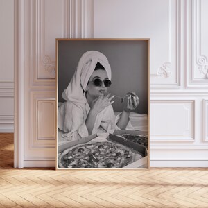 Pu&ograve; includere: Stampa in bianco e nero incorniciata di una persona con occhiali da sole e un asciugamano in testa, che mangia la pizza. Due scatole di pizza sono aperte su un letto. La stampa &egrave; esposta in una stanza con pareti bianche e pavimento in legno.