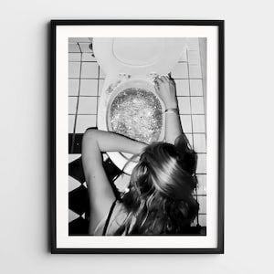 May include: Black and white framed print featuring a toilet with glitter inside. A person's arm and hair are visible, leaning towards the toilet. The background shows white and black tiled flooring. The image is a high-angle shot.