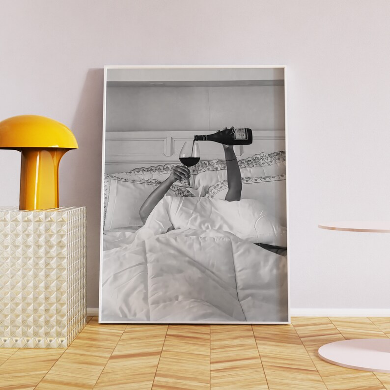 May include: Black and white framed print of a person pouring red wine into a glass while in bed. The image is a close-up, with the person's arms and the wine bottle visible. The bed is covered in white bedding.