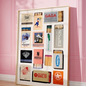 May include: A framed collection of vintage matchbooks, featuring various designs and logos. The matchbooks are arranged in rows and columns, showcasing brands like "GAGA PIZZERIA" and "ROCKY MOUNTAIN BAGEL CO." The frame is white, and the background is a pink wall.