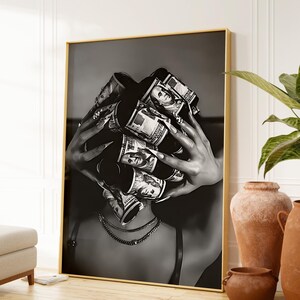 May include: Black and white framed art print featuring a person with rolled-up US dollar bills covering their face. The person is wearing a necklace and a bra. The artwork is displayed in a gold frame.
