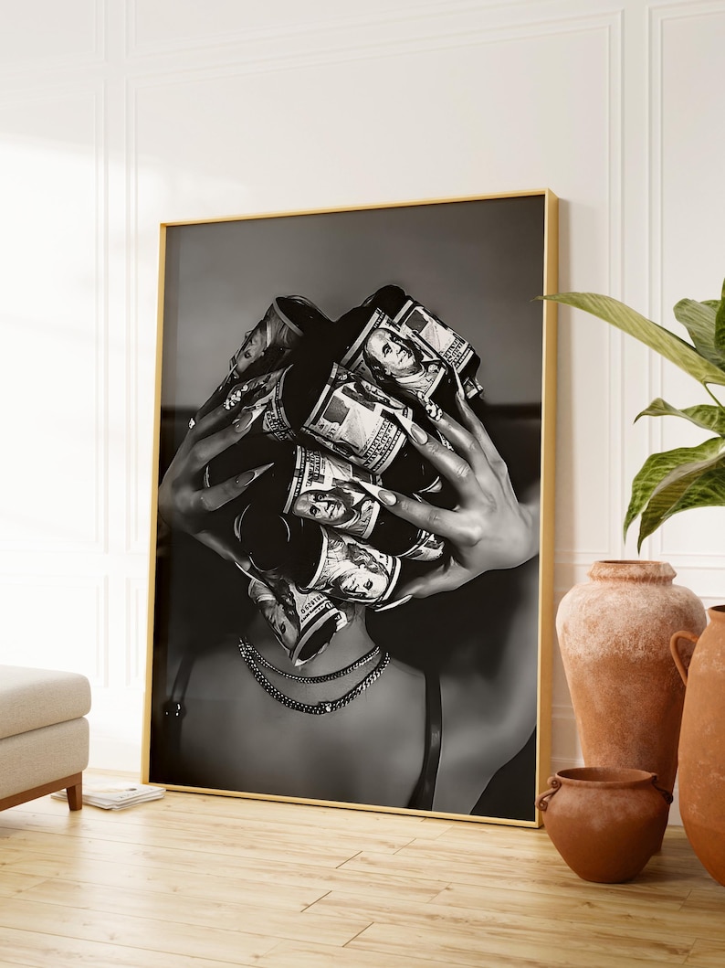 May include: Black and white framed art print featuring a person with rolled-up US dollar bills covering their face. The person is wearing a necklace and a bra. The artwork is displayed in a gold frame.
