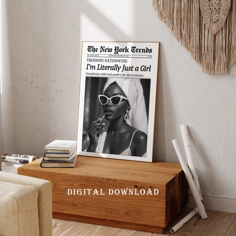 May include: A framed black and white print featuring a woman wearing sunglasses and a towel, applying lipstick. The print reads "The New York Trends" and "I'm Literally Just a Girl." The print is on a wooden table.