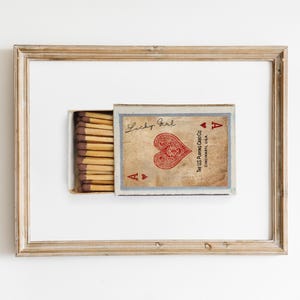 Puede incluir: Una caja de cerillas vintage enmarcada con un corazón rojo y el texto "Lucky Girl" y "The U.S. Playing Card Co. Cincinnati, U.S.A." La caja de cerillas está etiquetada con el As de Corazones.