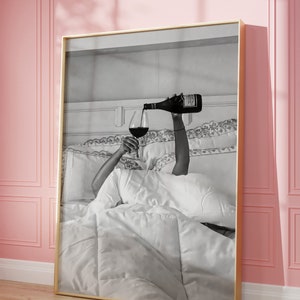 May include: Black and white framed art print. The image depicts a person pouring red wine into a glass while in bed. The bed has white linens and pillows. The frame is gold.