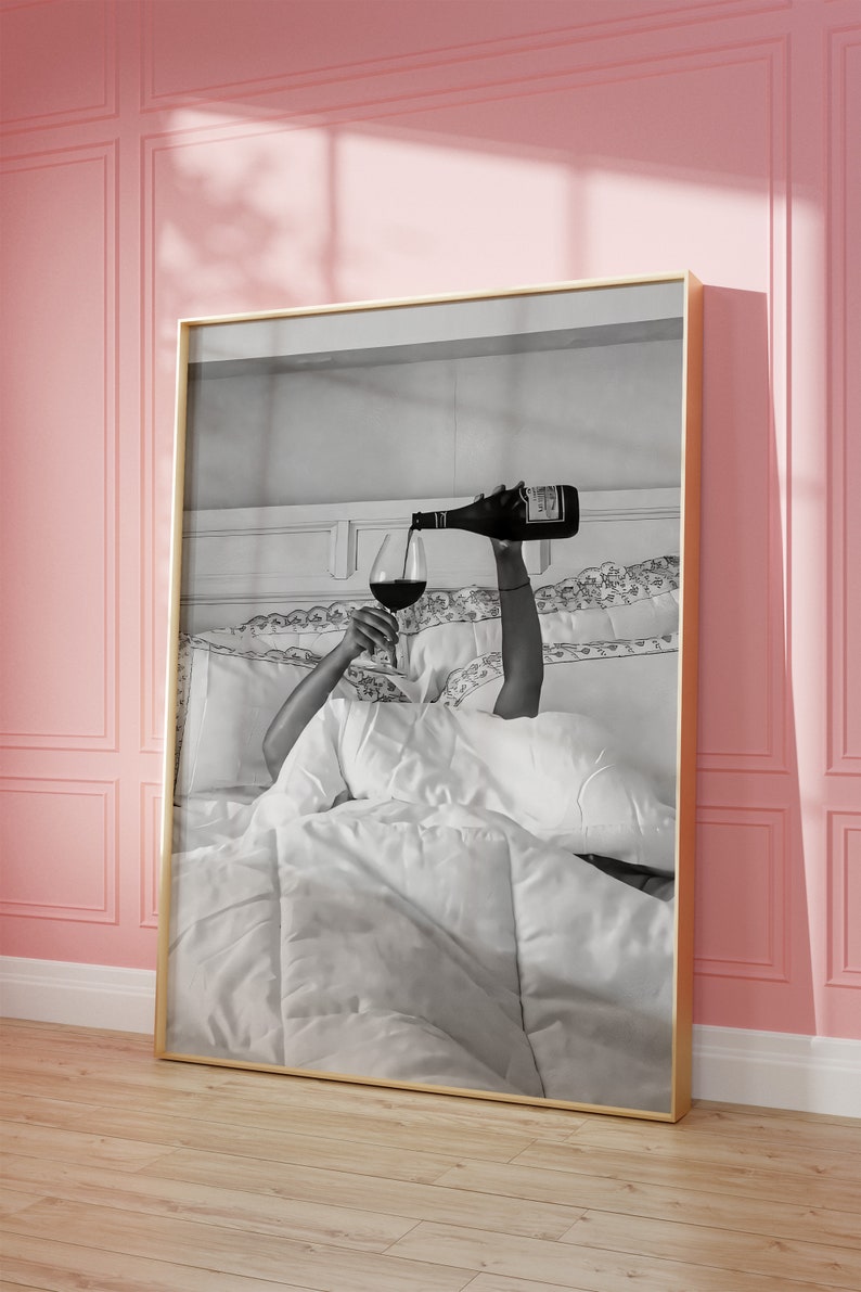 May include: Black and white framed art print. The image depicts a person pouring red wine into a glass while in bed. The bed has white linens and pillows. The frame is gold.