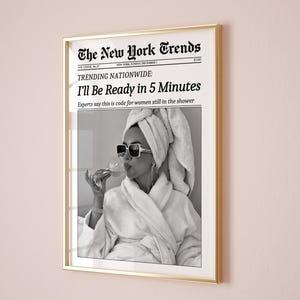 K&ouml;nnte beinhalten: Gerahmter Druck mit goldenem Rahmen. Der Druck ist eine gef&auml;lschte Zeitung, "The New York Trends", mit der Schlagzeile "I'll Be Ready in 5 Minutes". Das Bild ist ein Schwarz-Wei&szlig;-Foto einer Frau in Bademantel und Sonnenbrille.