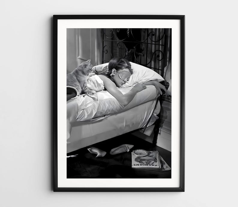 May include: A black and white photo of a woman sleeping in a bed with a cat. The woman is wearing a sleep mask and is lying on her side. The cat is curled up on the bed next to her. The photo is framed in a black frame.