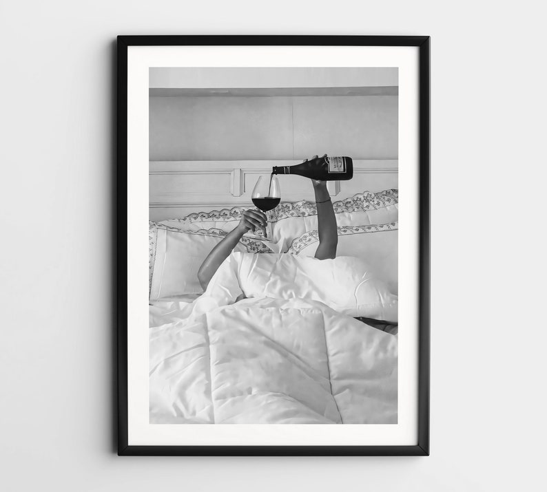 May include: Black and white framed print of a person pouring red wine into a glass while in bed. The person is under white bedding, with only their arms visible. The wine bottle is dark, and the glass is filled with red wine.
