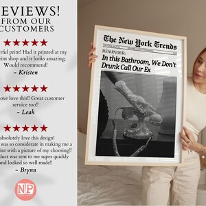 May include: A framed black and white print with the text "The New York Trends" and "In this Bathroom, We Don't Drunk Call Our Ex". The print features a vintage rotary phone with a hand reaching towards it. The print is held by a person wearing a white top and beige trousers.