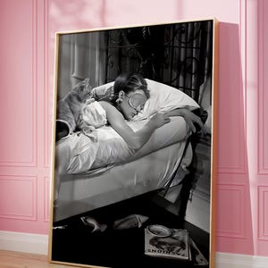May include: Black and white photograph of a woman sleeping in bed with a cat. The woman is wearing a sleep mask and has her arm around the cat. The bed is made with white sheets and a white pillow. The woman is wearing a white shirt and the cat is lying on top of the sheets. The photo is framed in a gold frame. The text "FASHIONS" is visible on a magazine on the floor.