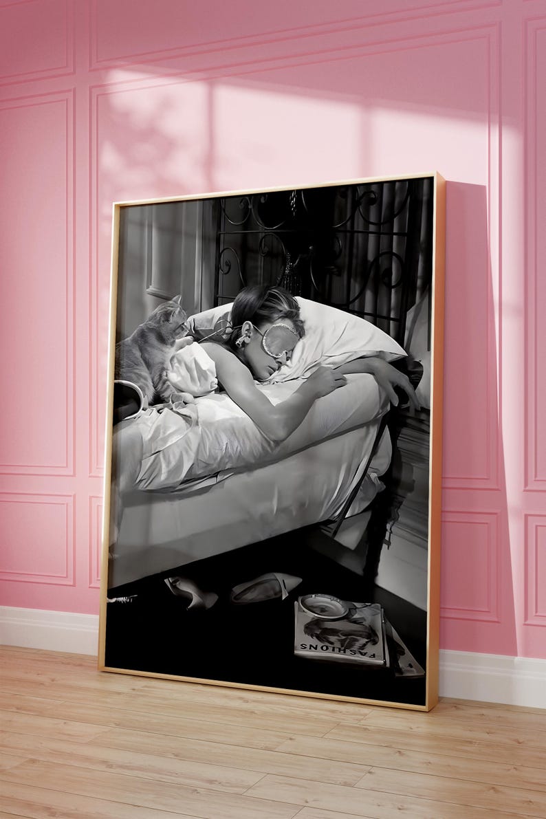May include: Black and white photograph of a woman sleeping in bed with a cat. The woman is wearing a sleep mask and has her arm around the cat. The bed is made with white sheets and a white pillow. The woman is wearing a white shirt and the cat is lying on top of the sheets. The photo is framed in a gold frame. The text "FASHIONS" is visible on a magazine on the floor.
