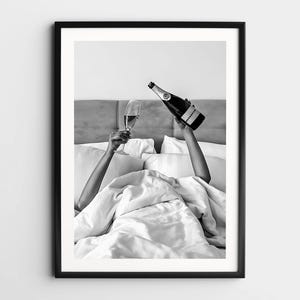 May include: A black and white framed wall art print featuring two arms raised from beneath white bed sheets. One hand holds a tall champagne flute, while the other hand holds a bottle of champagne tilted as if pouring into the glass. The scene is set against a headboard.