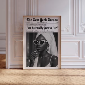 May include: A framed black and white print featuring a woman wearing sunglasses and a towel on her head, applying lipstick. The print is designed to look like a newspaper, with the headline "The New York Trends" and the text "I'm Literally Just a Girl."