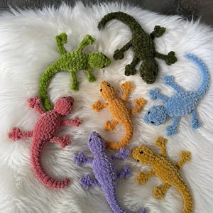 May include: A collection of six plush lizard toys made from soft, textured yarn. The lizards are colored in bright green, dark olive green, light blue, pink, orange, purple, and yellow, each featuring small black eyes and four splayed legs.