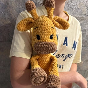 May include: A crocheted giraffe toy made with brown and yellow yarn. The giraffe has large brown eyes and a brown nose. The toy is being held by a person wearing a yellow shirt.