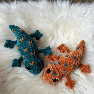 May include: Two plush lizard toys made of soft, textured yarn. One lizard is dark teal with orange spots, and the other is orange with dark teal spots. Both feature large, smooth, black plastic eyes and four splayed legs with distinct toes.