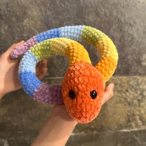 Puede incluir: Un juguete de serpiente de crochet arcoíris con una cabeza naranja brillante y ojos negros.