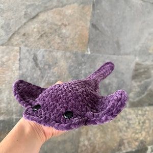 May include: A purple crocheted stingray plush toy with black eyes. The toy is being held in a person's hand.