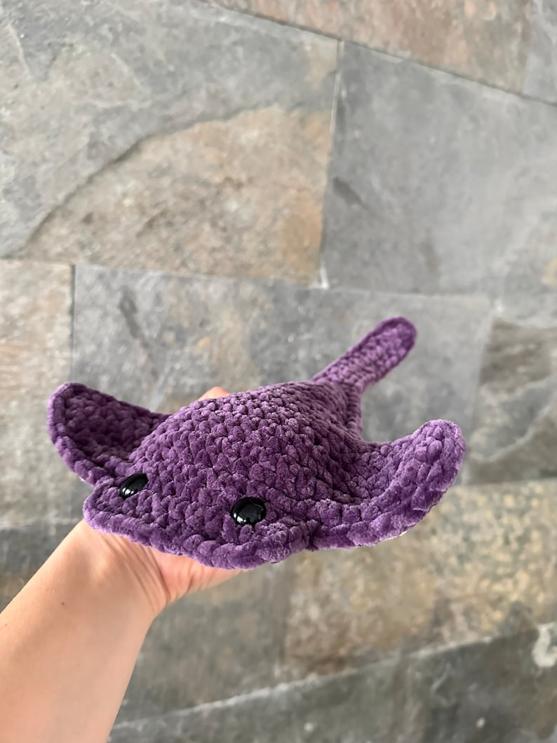 May include: A purple crocheted stingray plush toy with black eyes. The toy is being held in a person's hand.