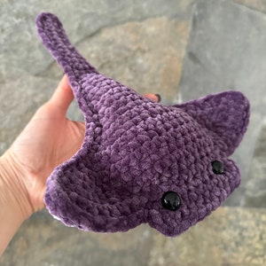 May include: A plush purple crocheted stingray with black eyes. The stingray is held in a hand.