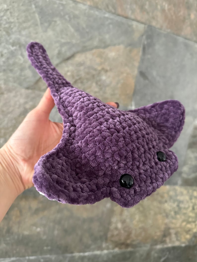 May include: A plush purple crocheted stingray with black eyes. The stingray is held in a hand.