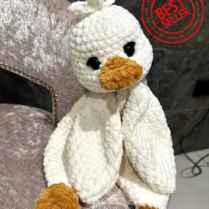 May include: White crocheted plush duck toy with brown felt beak and eyes. The duck is sitting on a chair with a plush fabric.