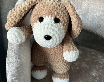 May include: A crocheted plush dog toy with brown and white fur. The dog is standing on a gray chair.