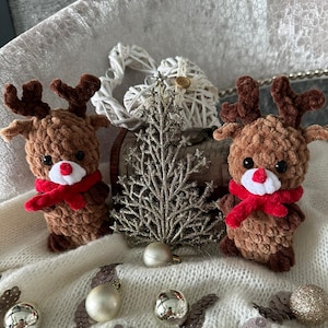May include: Two handmade brown crochet reindeer with red scarves and brown antlers. A silver Christmas tree and ornaments are in the background. The reindeer have white noses and black eyes. The scene is set on a white, textured surface.